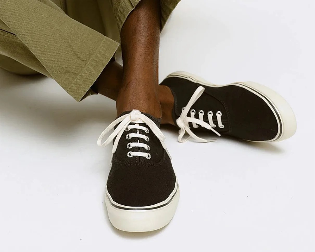 Black canvas sneakers with white laces and thick white soles worn with olive green pants.