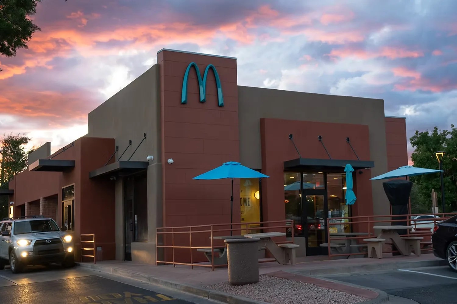 Fast-food restaurant building with a blue McDonald's logo, outdoor seating with blue umbrellas, and cars in the parking lot at sunset.