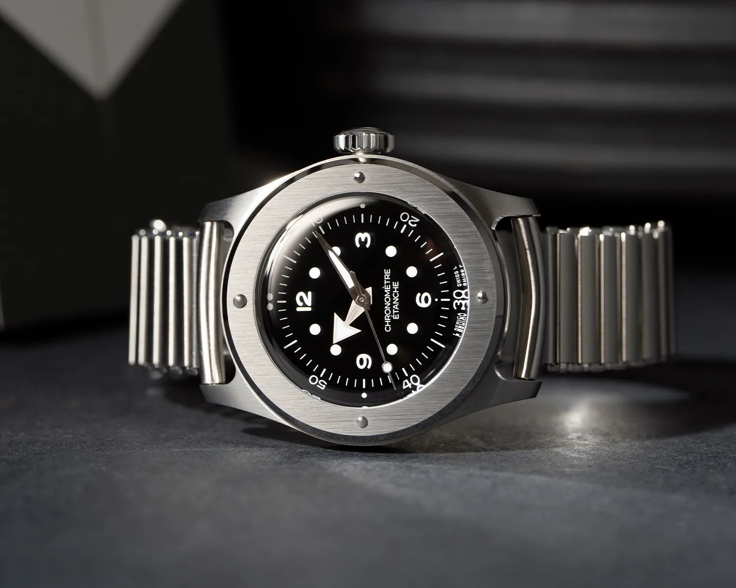 Stainless steel wristwatch with black dial, white numerals, and a metal expandable bracelet on a dark surface.