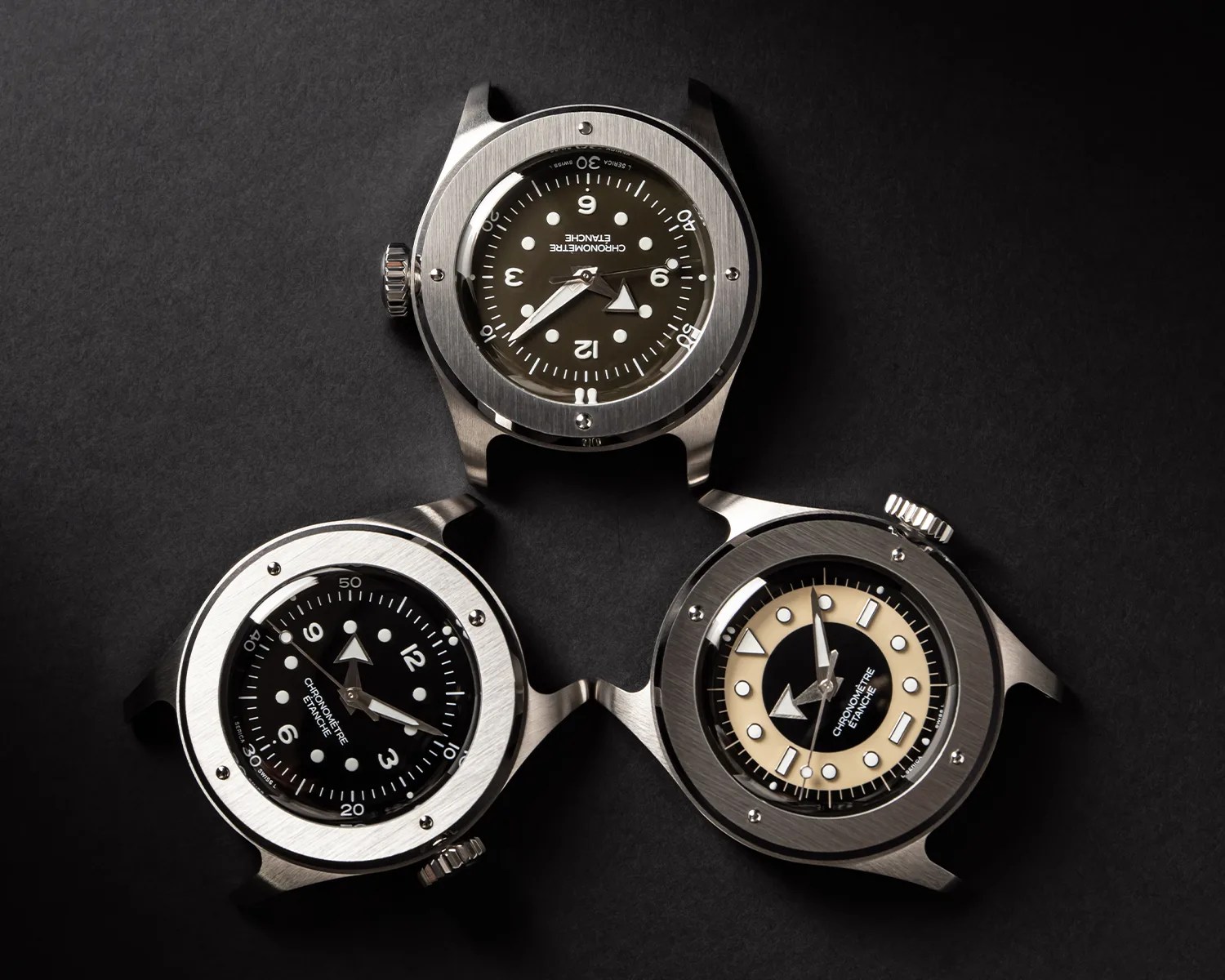 Three stainless steel watch cases with black and beige dials arranged in a triangular pattern on a black surface.