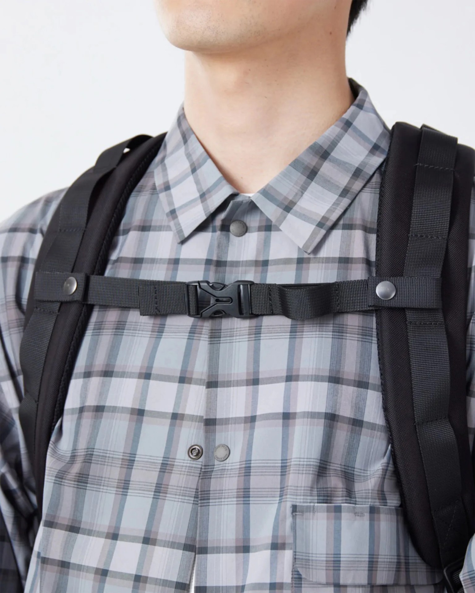Close-up of a person wearing a gray plaid button-up shirt with a black backpack chest strap fastened.