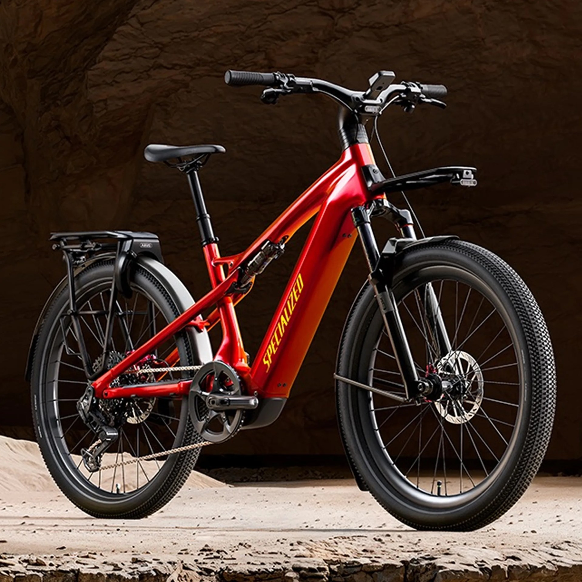 Red Specialized electric mountain bike with black tires, fenders, and rear rack on sandy terrain.