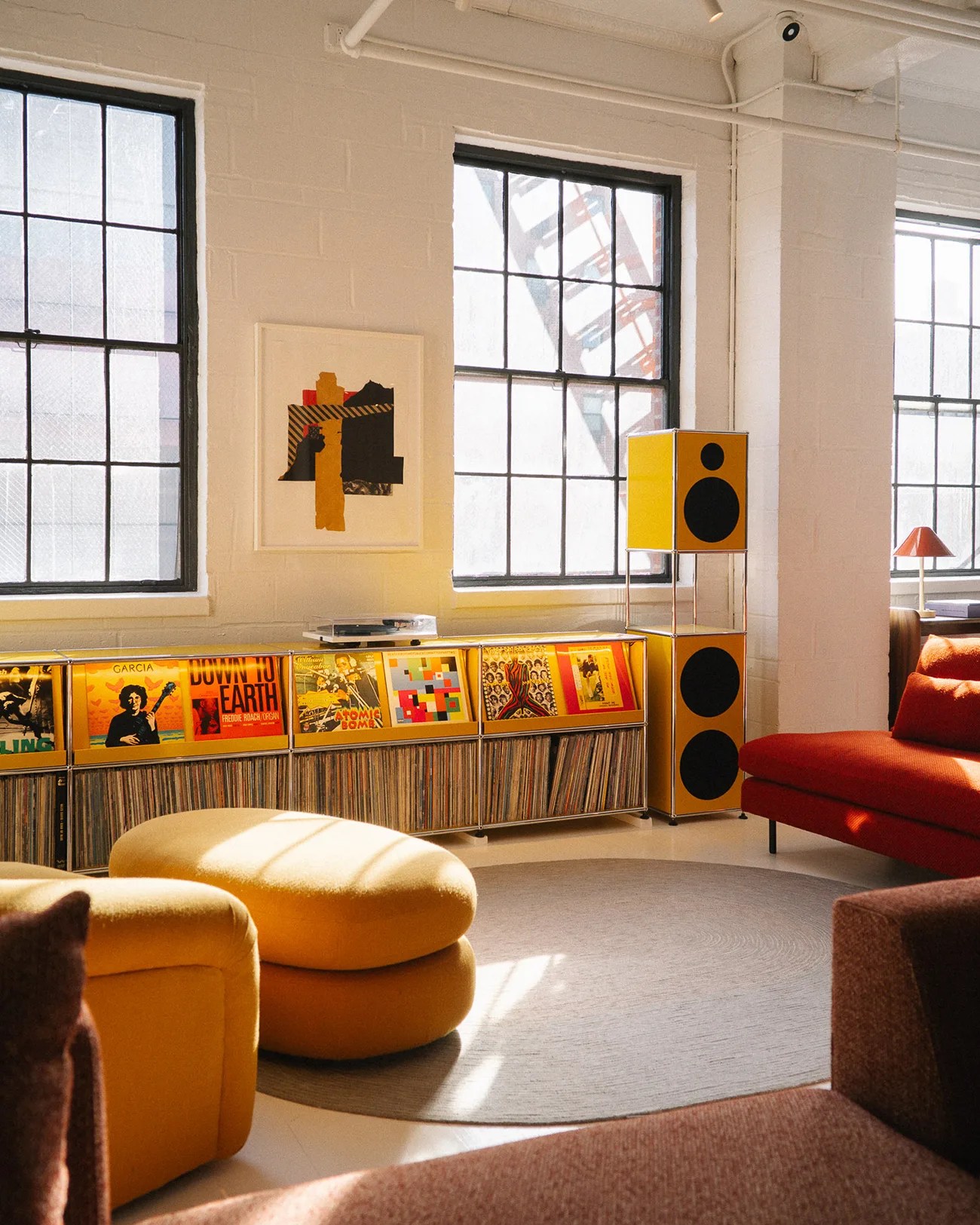 Sunlit modern living room with yellow ottomans, red sofa, yellow speakers, and a vinyl record collection under large windows.