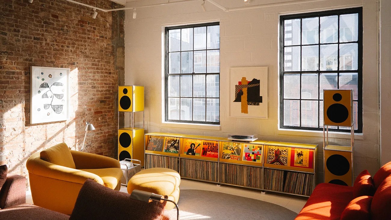 Sunlit living room with exposed brick wall, yellow armchair and ottoman, red sofa, and yellow speakers with vinyl records.