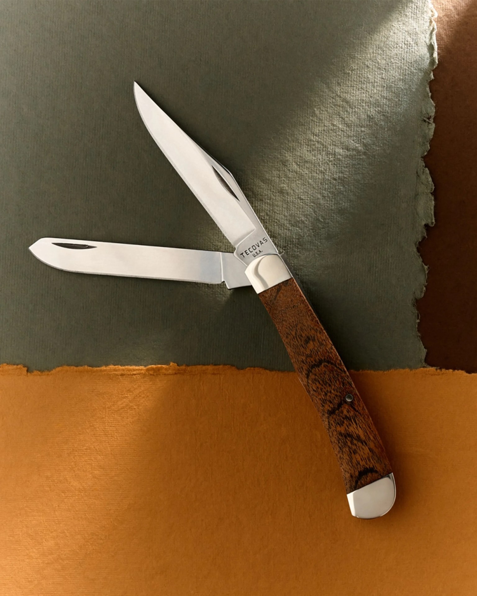 Folding pocket knife with two stainless steel blades and a wooden handle on a textured green and brown surface.
