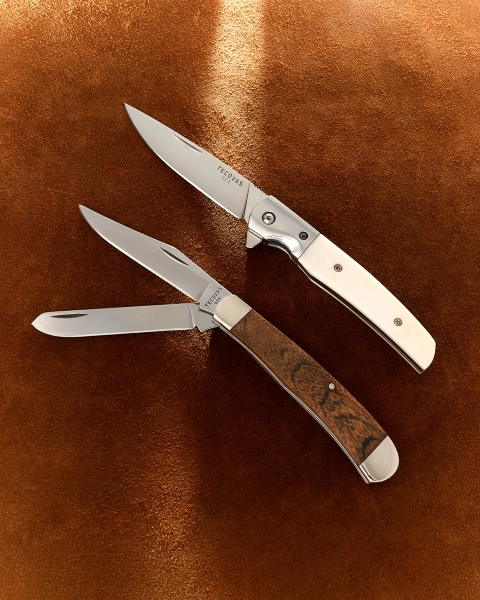 Three folding knives with stainless steel blades and handles in white, wood, and metal finishes on a brown textured surface.
