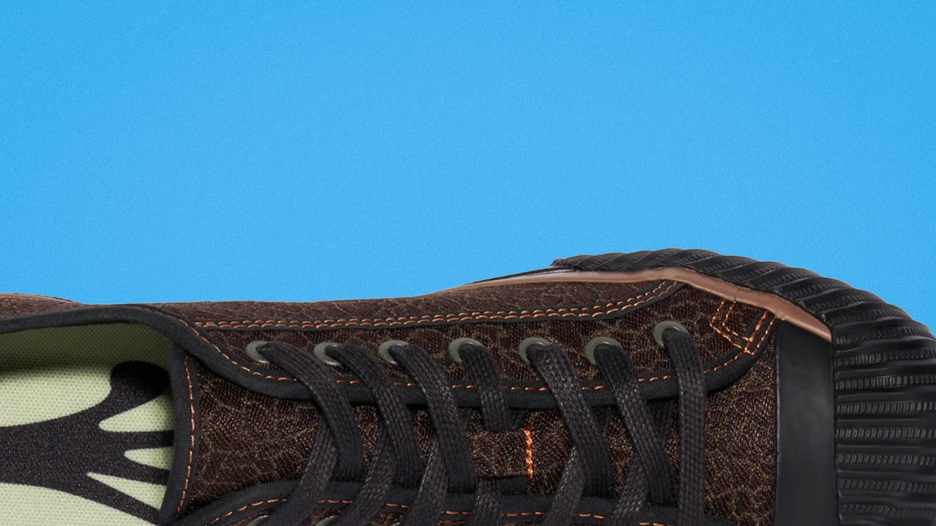 Close-up of a brown textured sneaker with black laces, orange stitching, and a black rubber toe cap against a blue background.