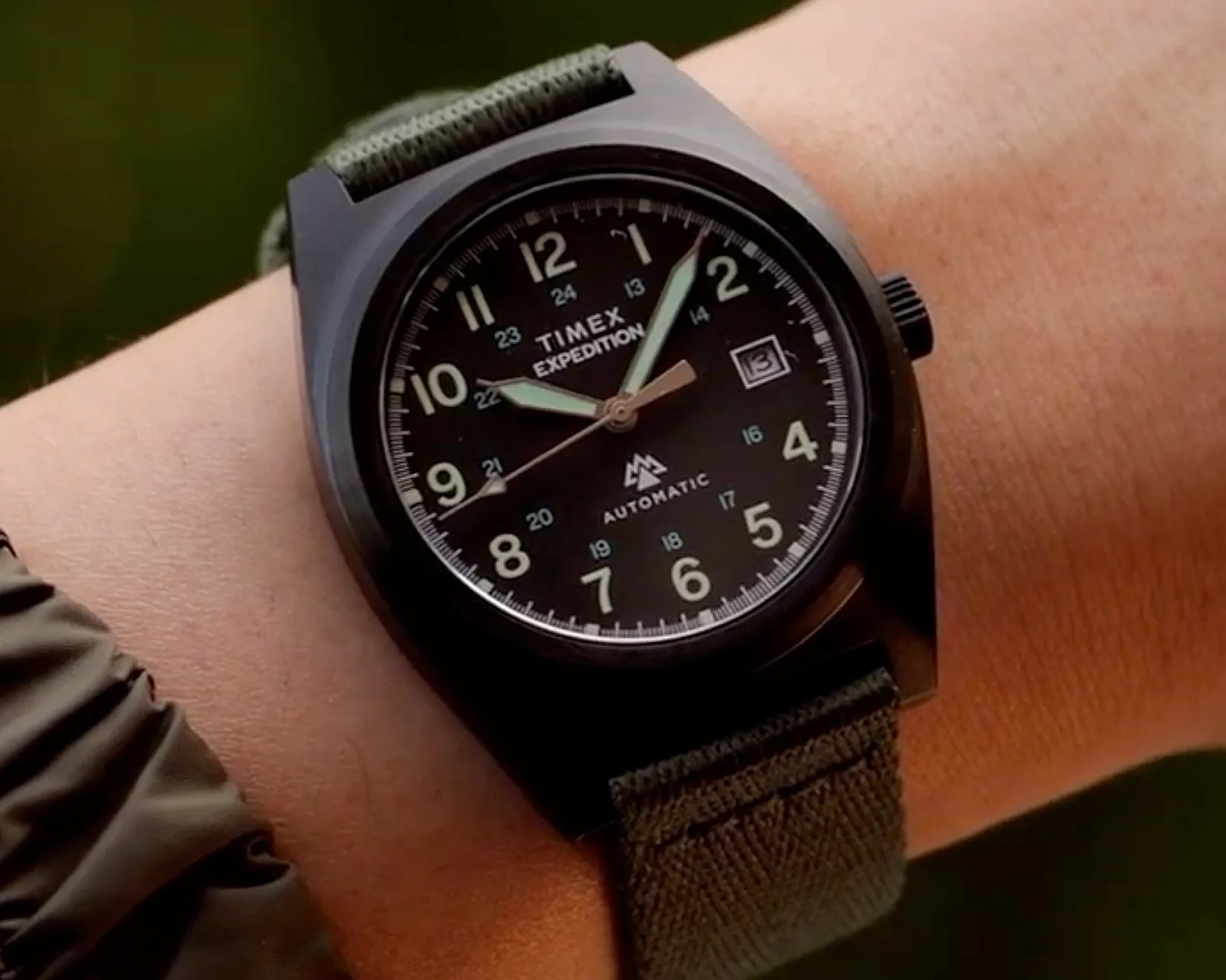 Black Timex Expedition wristwatch with green fabric strap and luminous hands on a wrist.