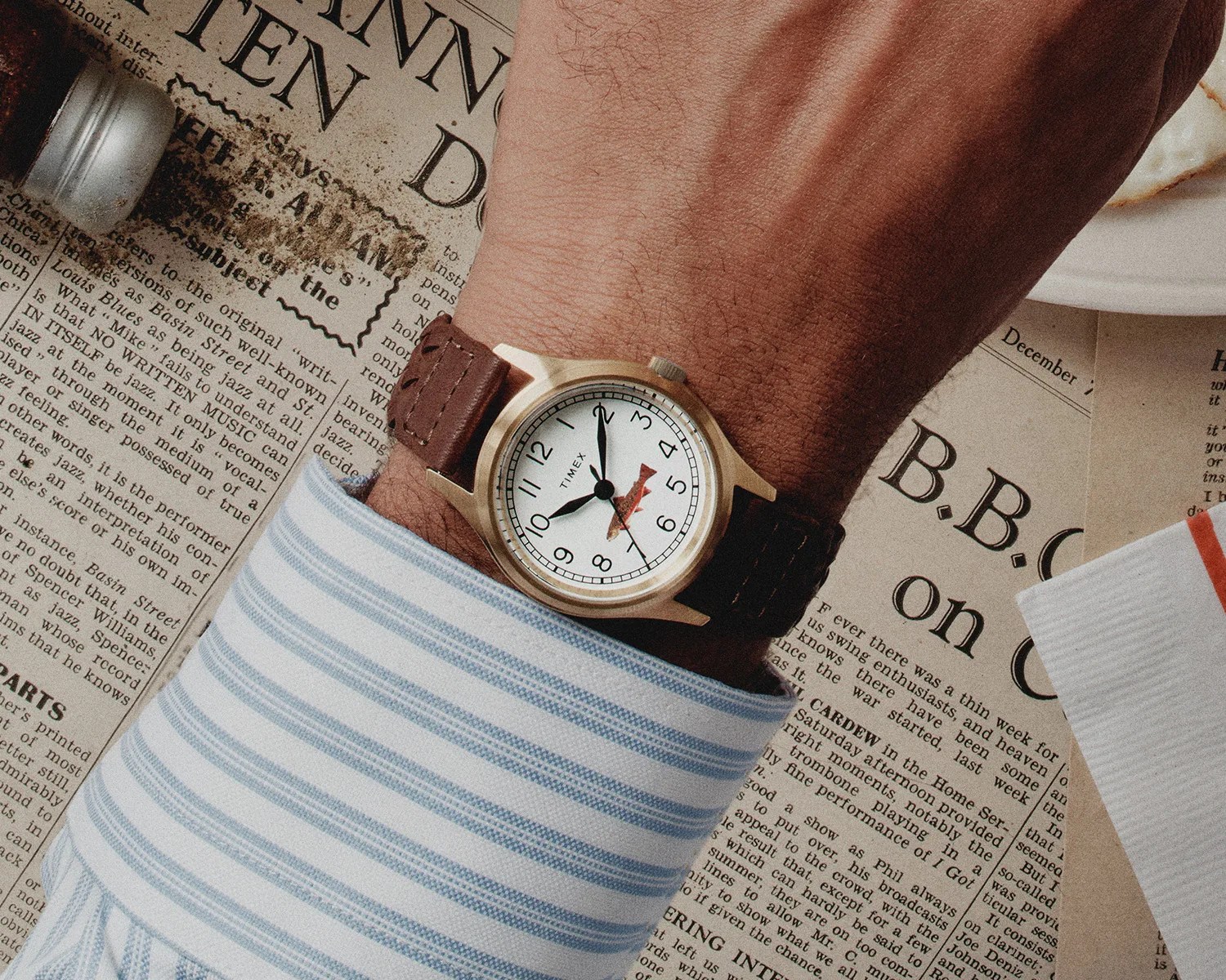 Wristwatch with a white dial, black numerals, and a brown leather strap worn on a wrist over a blue-striped shirt cuff.