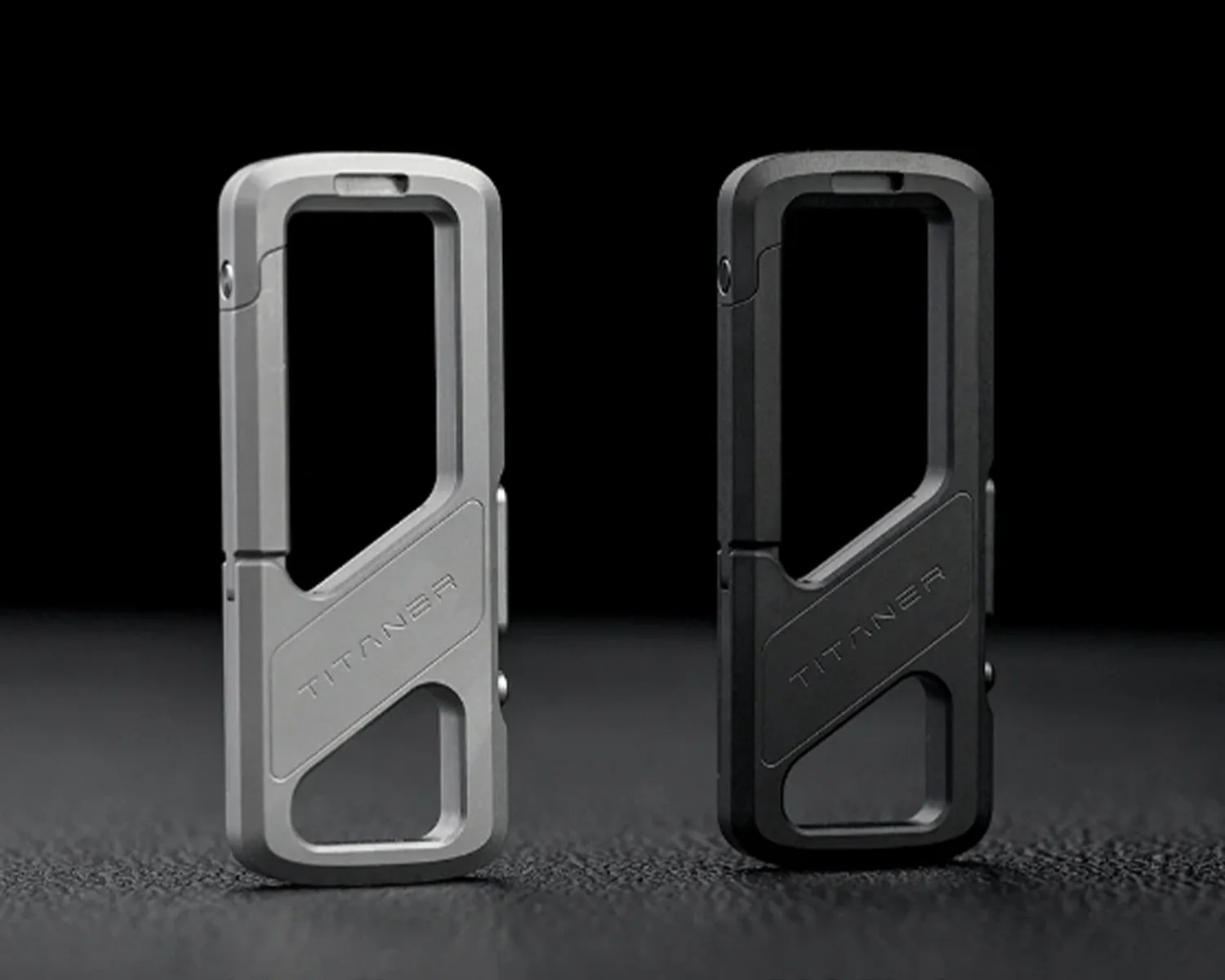Two rectangular metal carabiner clips, one silver and one black, each engraved with "TITANER" on a textured dark surface.