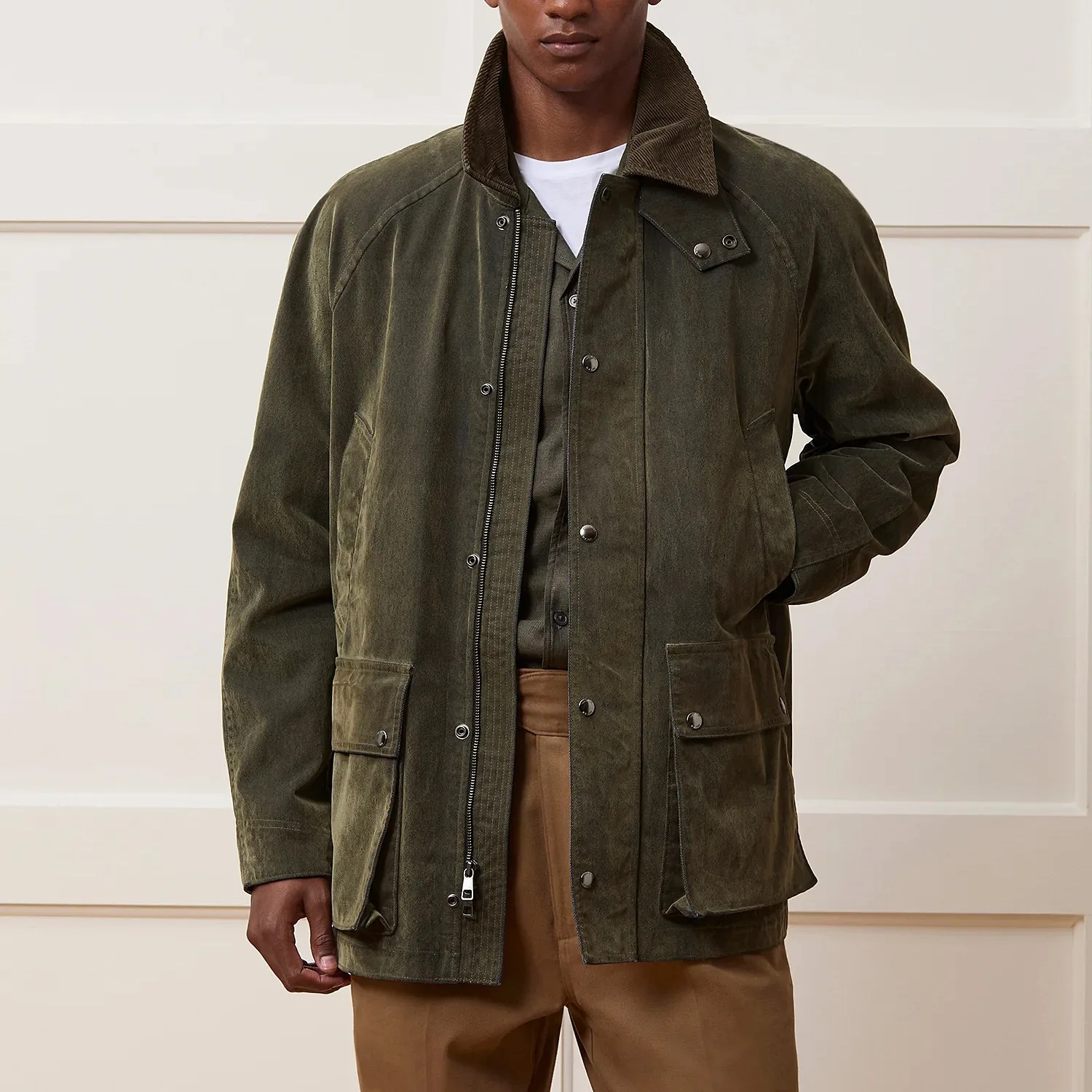 Olive green waxed cotton jacket with corduroy collar and flap pockets worn over brown pants.