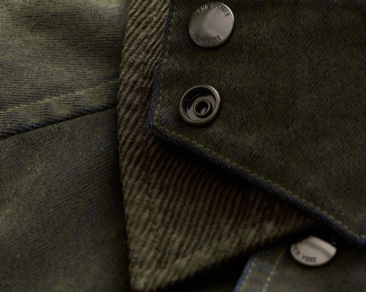 Close-up of dark green corduroy fabric with metal snap buttons, one engraved with "TODD SNYDER NEW YORK.