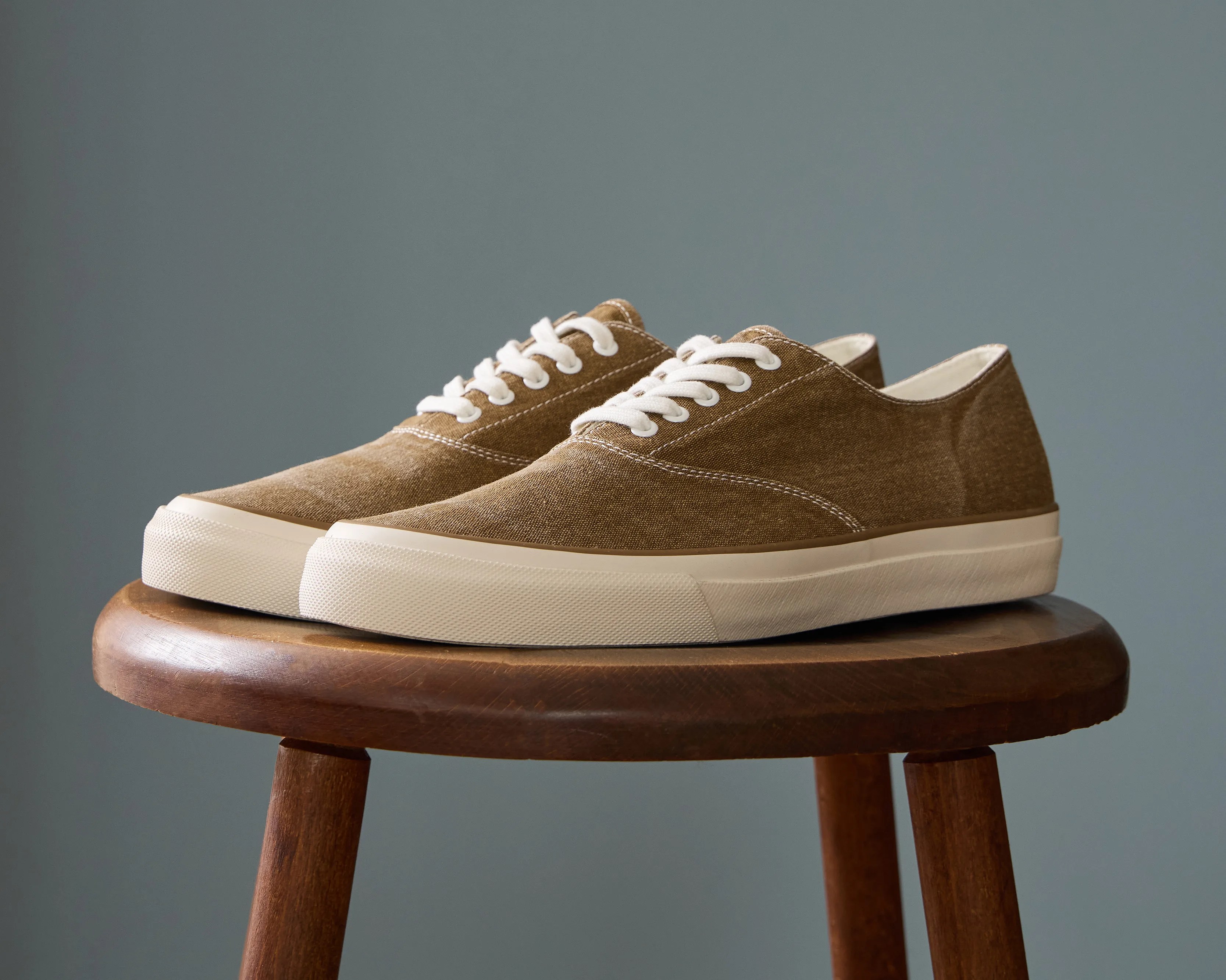 Pair of brown canvas sneakers with white laces and thick white rubber soles on a wooden stool.