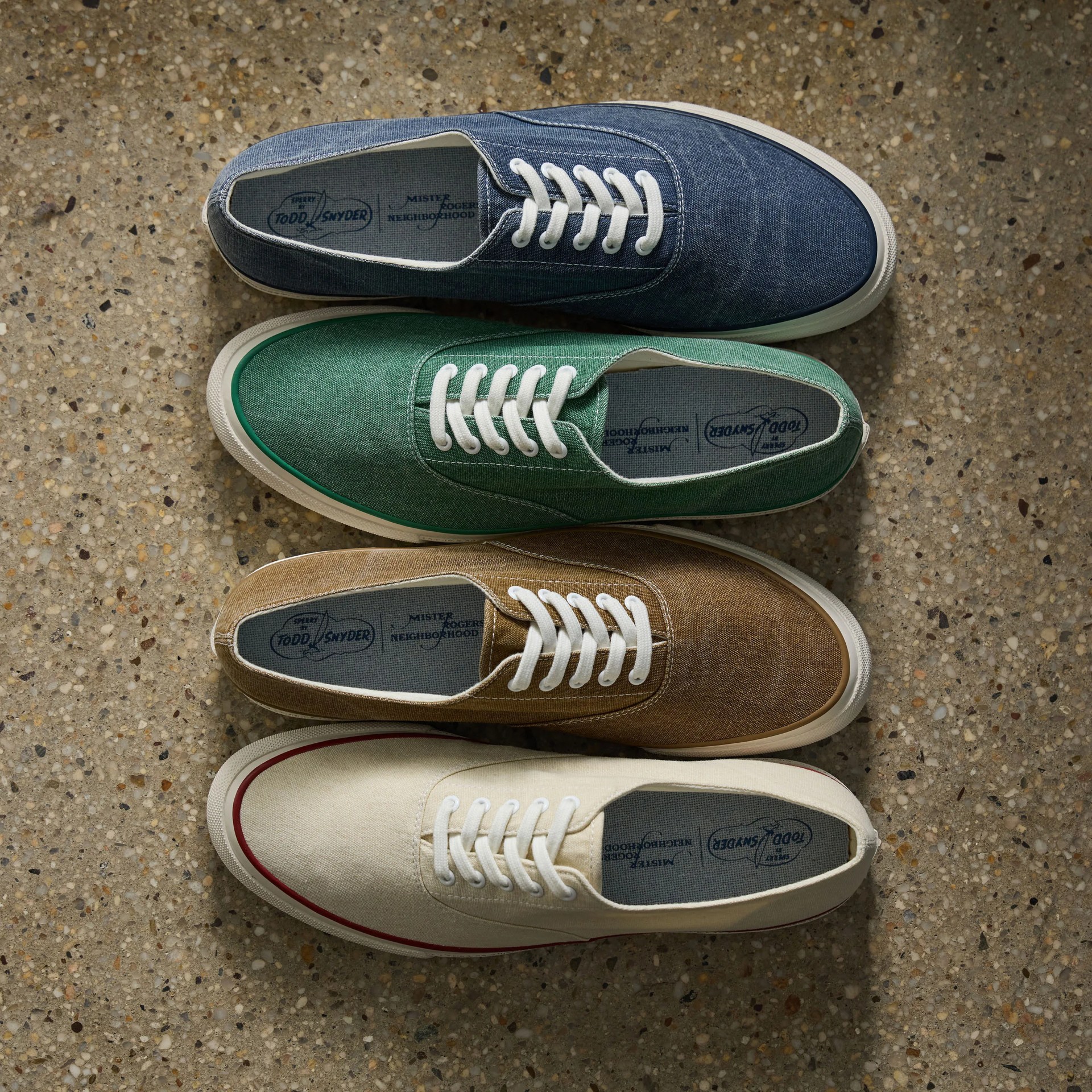 Four pairs of canvas sneakers in blue, green, brown, and off-white with white laces on a terrazzo floor.