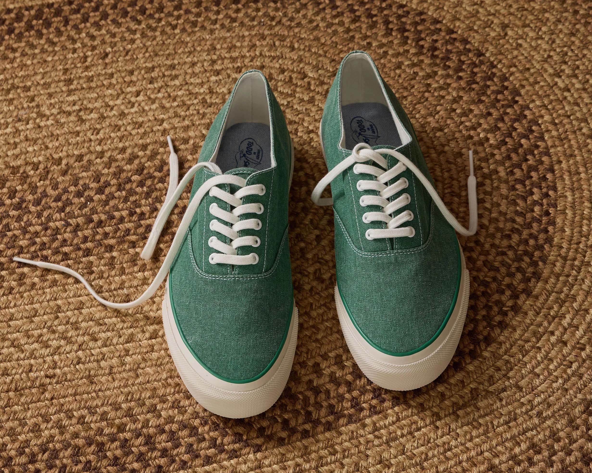 Pair of green canvas sneakers with white laces on a textured brown woven rug.