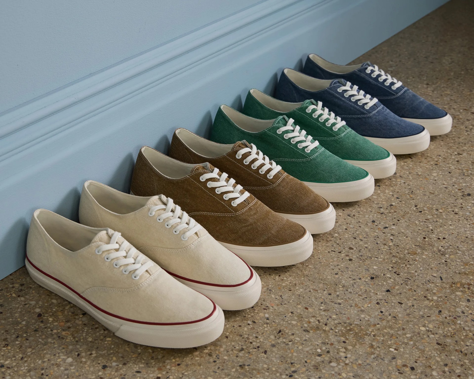 Row of canvas sneakers in cream, brown, green, and navy lined up against a blue wall on a terrazzo floor.