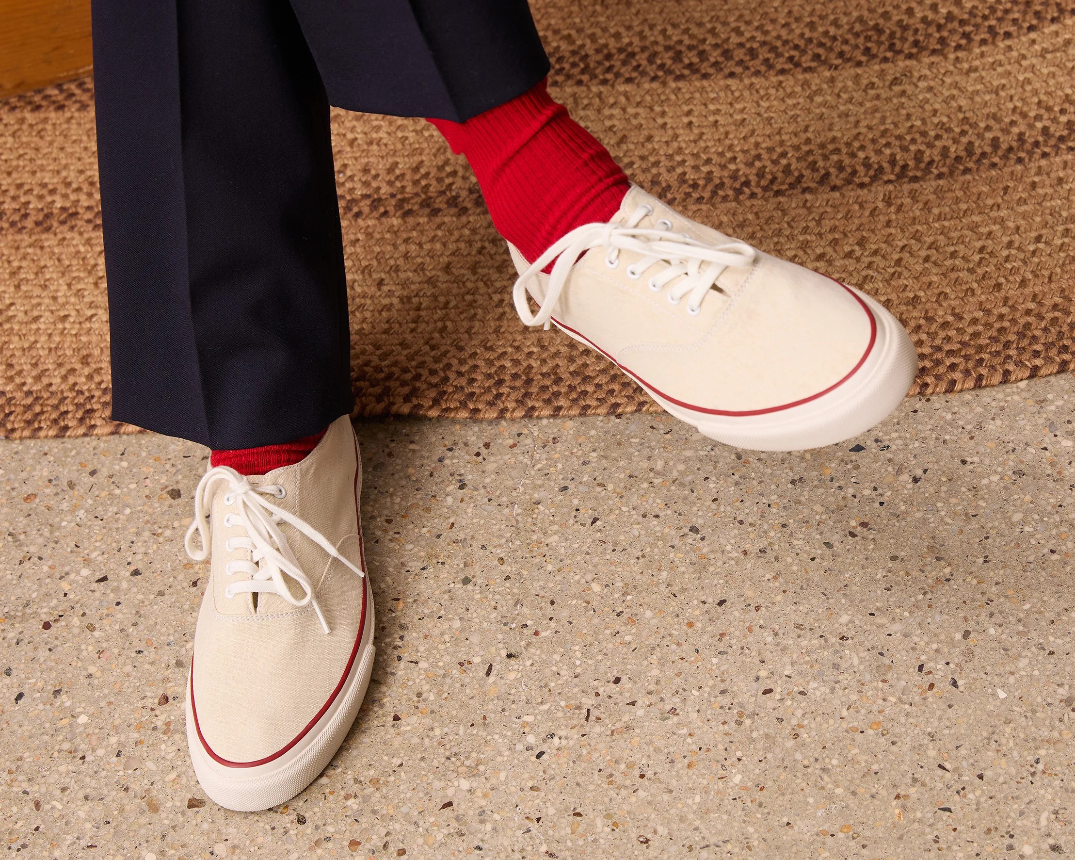 Off-white canvas sneakers with white laces and red trim worn with red socks and black pants.