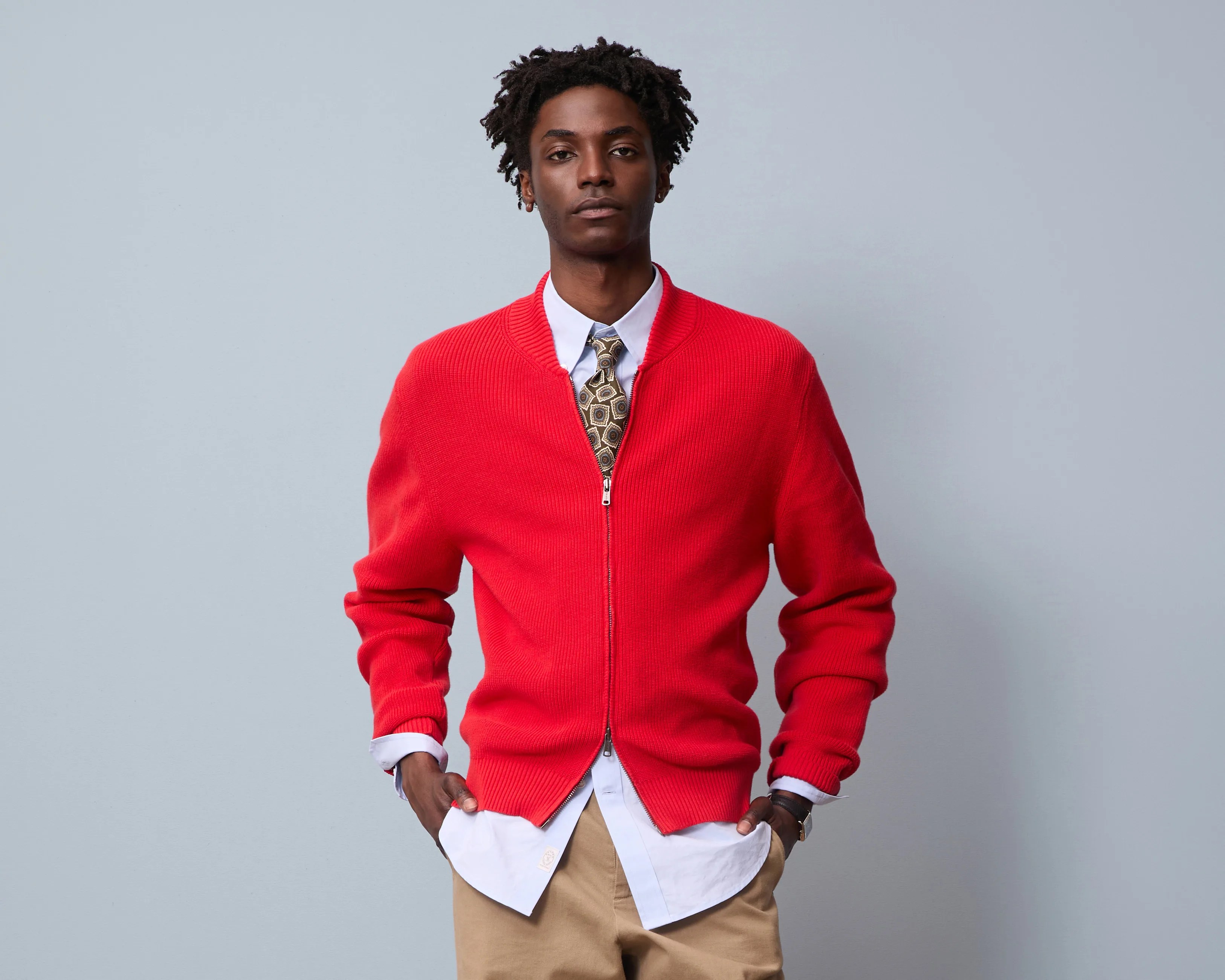 Man wearing a red zip-up sweater over a white shirt with a patterned tie and beige pants against a gray background.
