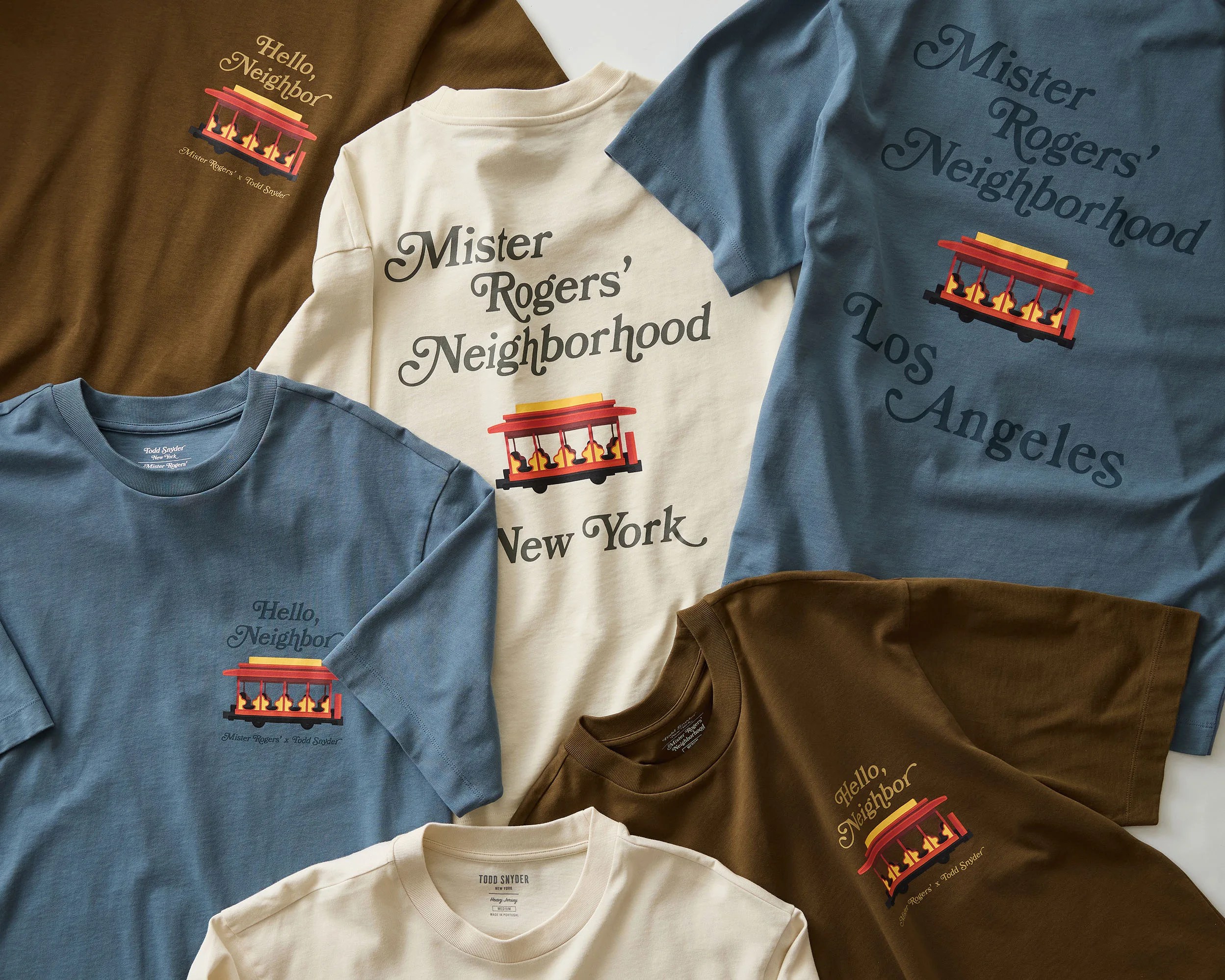Assorted brown, cream, and blue Mister Rogers' Neighborhood T-shirts with trolley graphic and text.