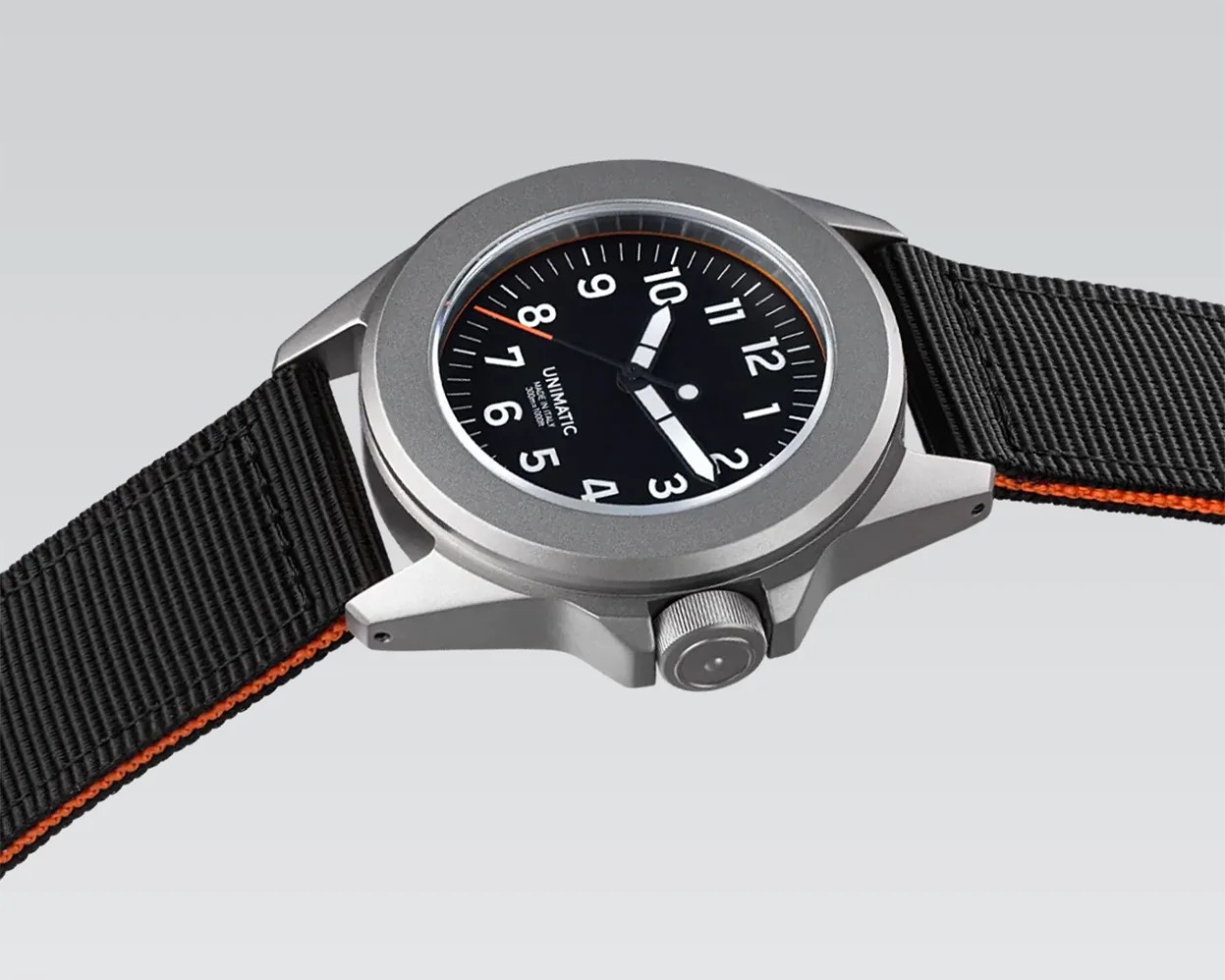 Silver Unimatic wristwatch with black dial, white numerals, and black fabric strap with orange edge.