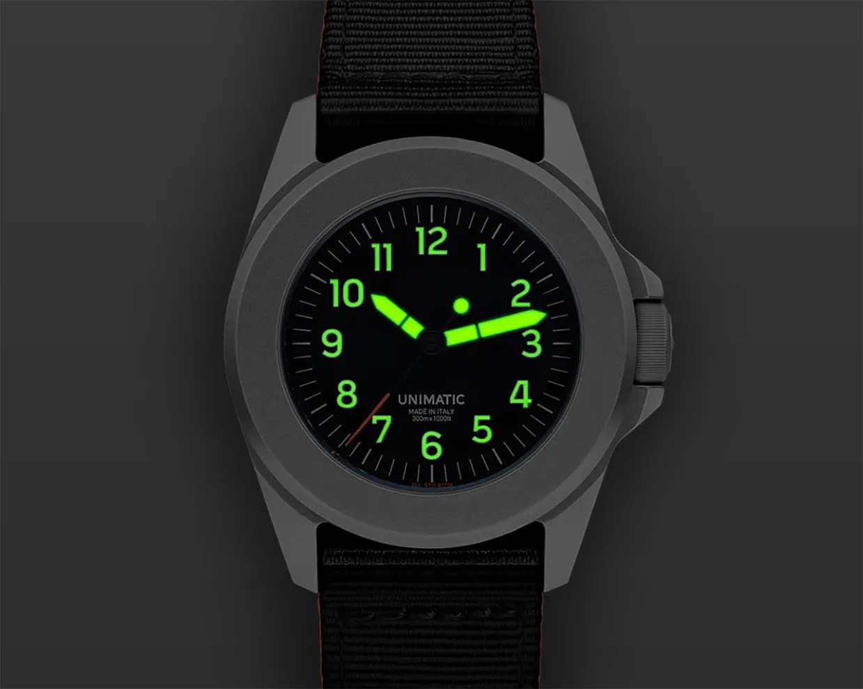 Matte gray Unimatic wristwatch with black dial and glowing green numerals and hands on a black fabric strap.