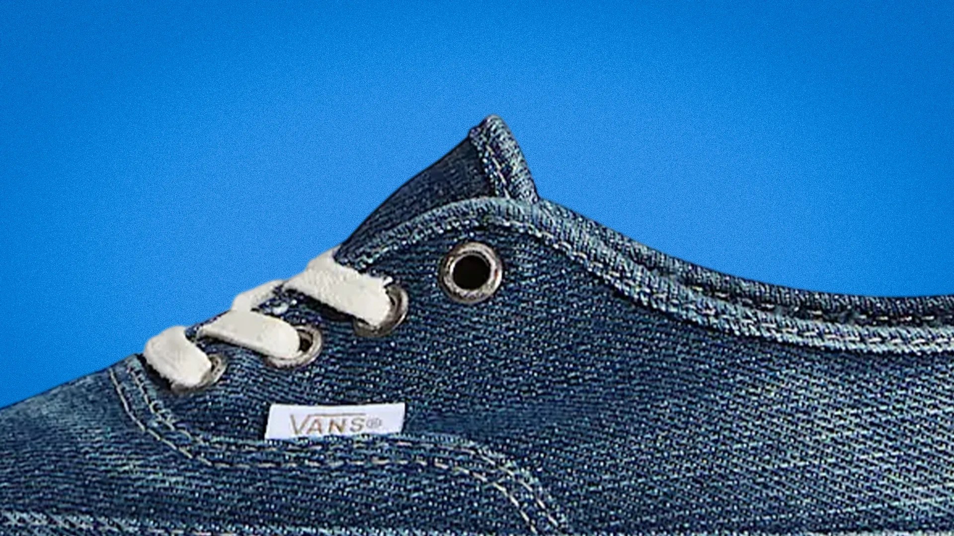 Close-up of a blue denim Vans sneaker with white laces and metal eyelets against a blue background.