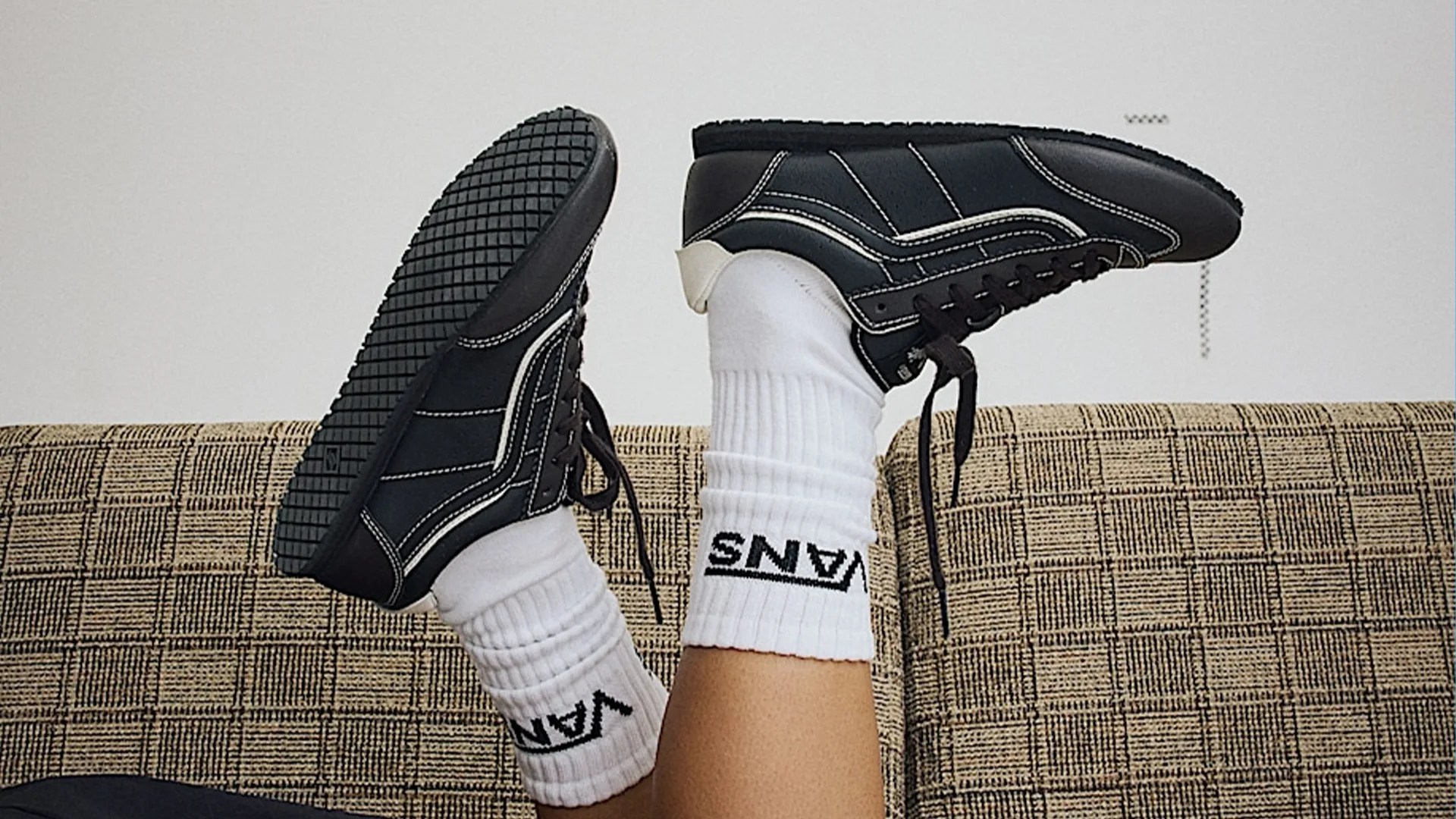 Black sneakers with white stitching worn with white Vans socks on a patterned beige couch.