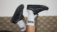 Black sneakers with white stitching worn with white Vans socks on a patterned beige couch.