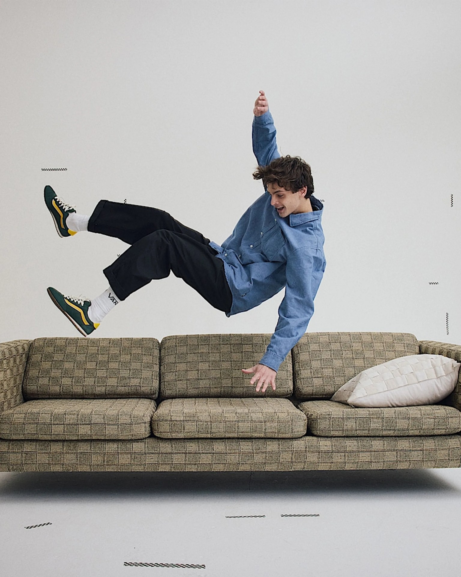 Young person in a blue jacket, black pants, and green sneakers mid-air above a brown plaid sofa with a white pillow.