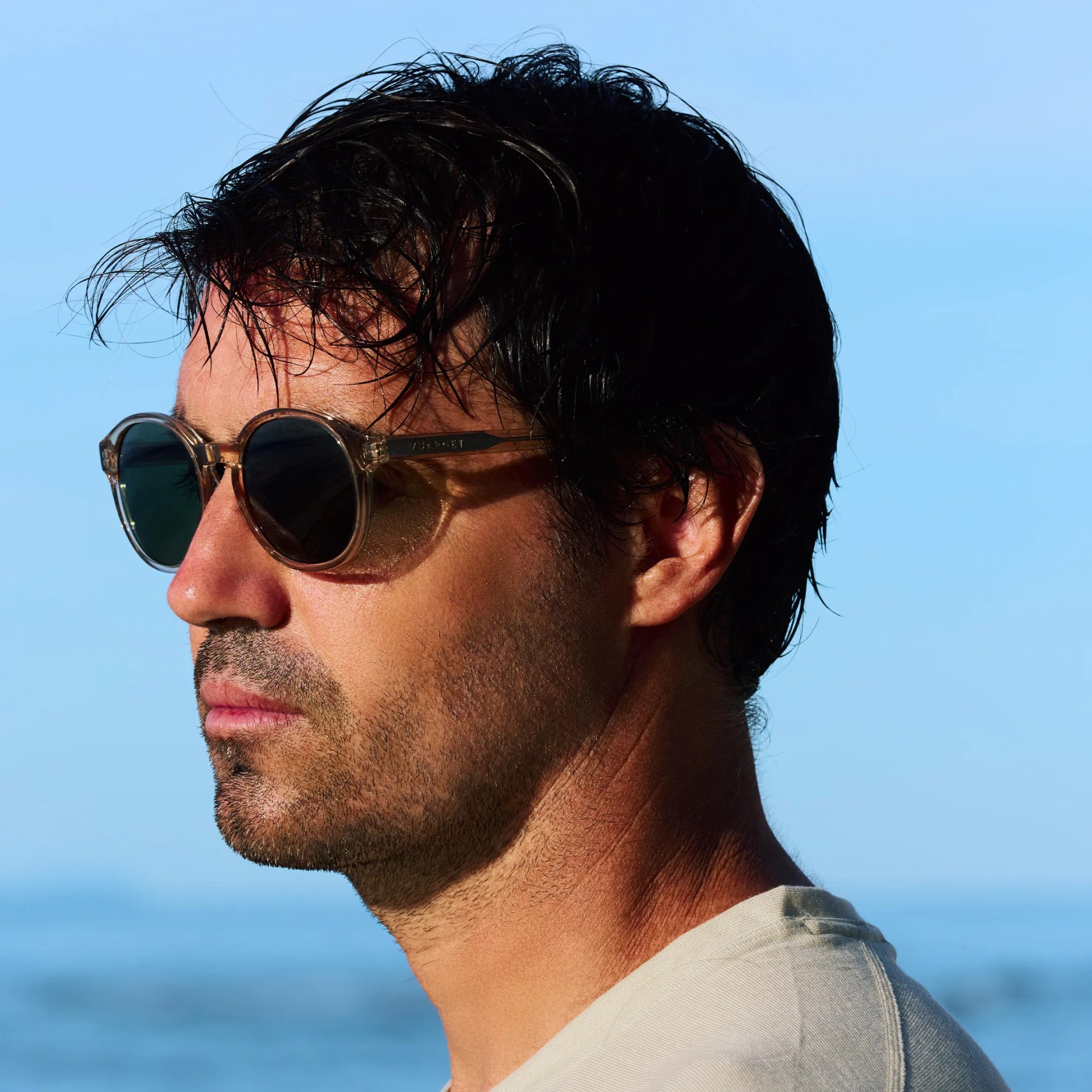 Close-up side profile of a man with wet black hair wearing round sunglasses against a clear blue sky and sea background.