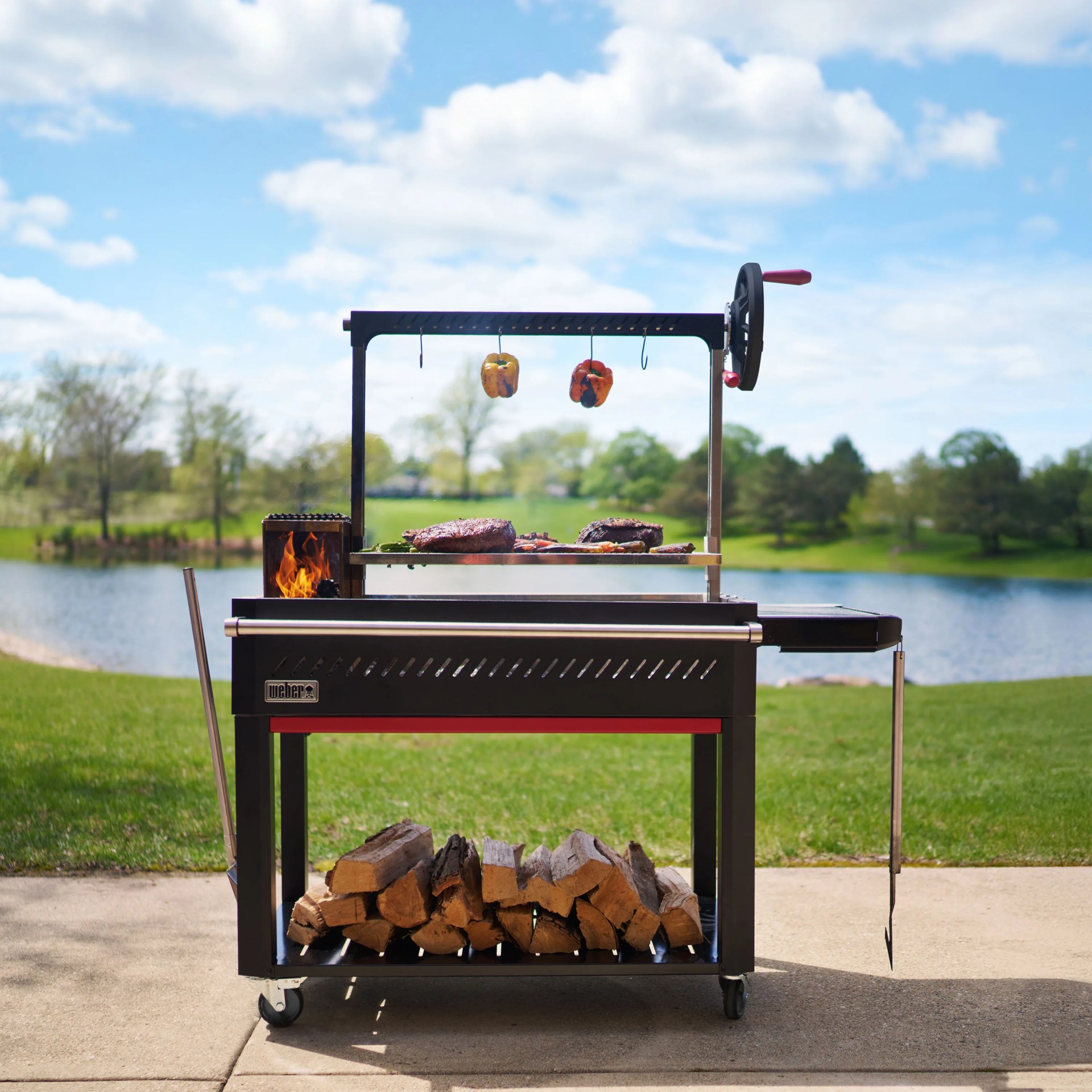 Black Weber grill with firebox on left, hanging grilled peppers, and stacked firewood underneath on wheels.