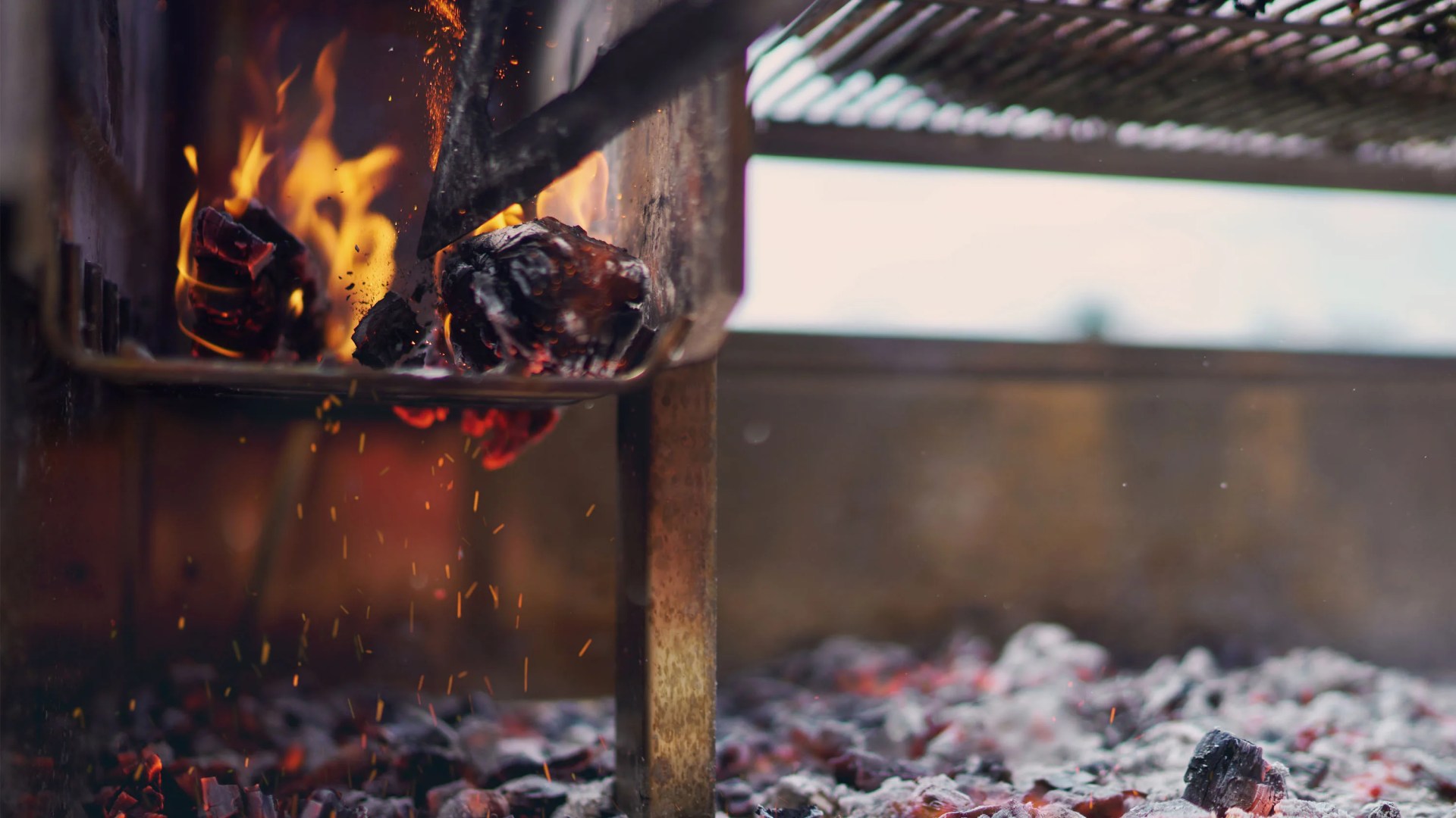 Close-up of glowing charcoal briquettes burning with orange flames and sparks inside a metal grill.