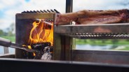 Flames burning charcoal in a metal grill with meat cooking on a rack above.