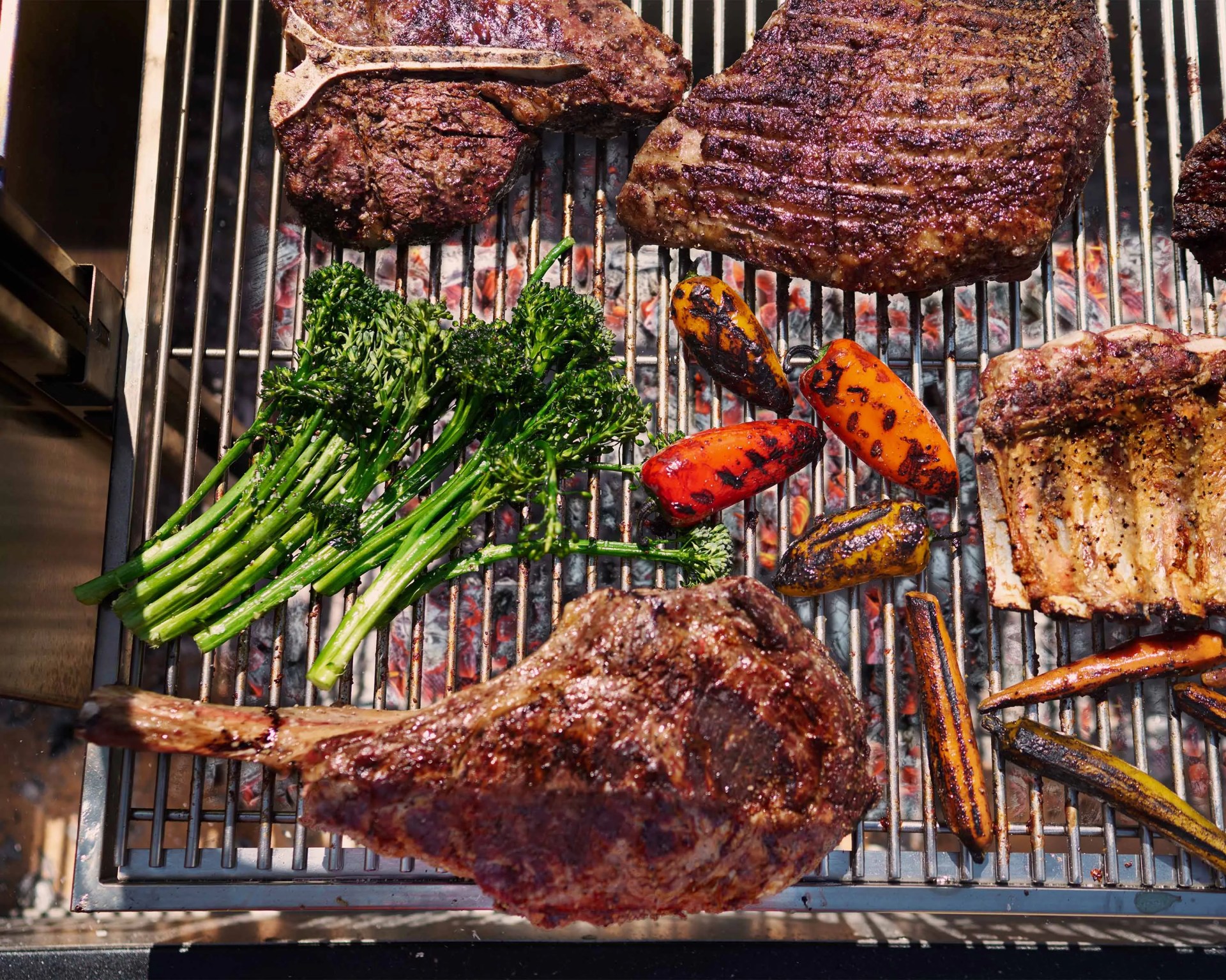 Grilled T-bone steaks, broccolini, mini bell peppers, and carrots on a barbecue grill over hot coals.
