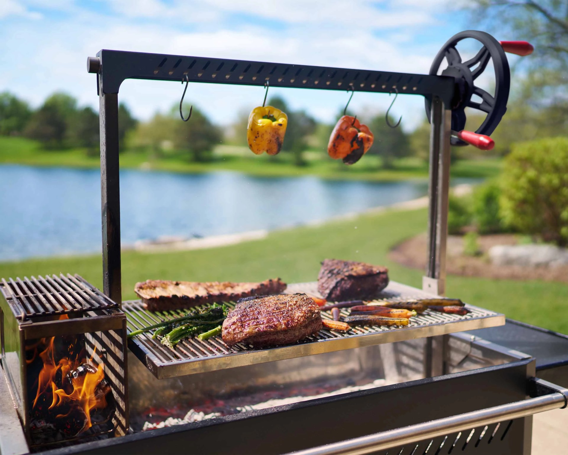 Outdoor grill with steaks, vegetables, and charred bell peppers hanging on hooks above, near a lake.
