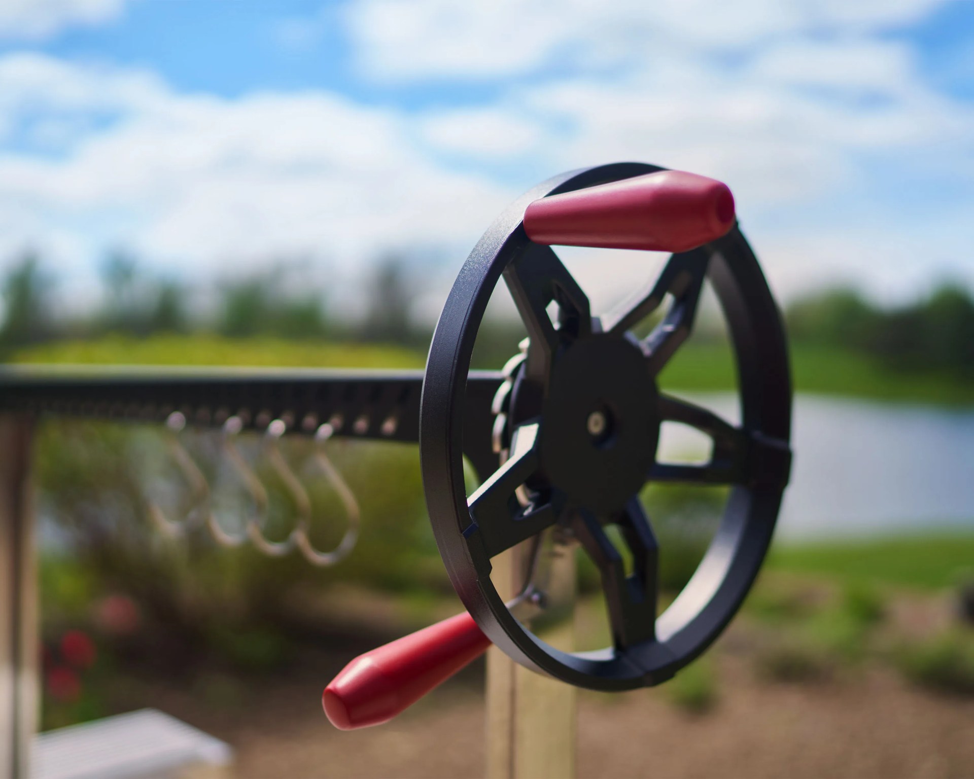 Close-up of a black metal wheel with red handles attached to a mechanism outdoors with blurred greenery and blue sky in the background.