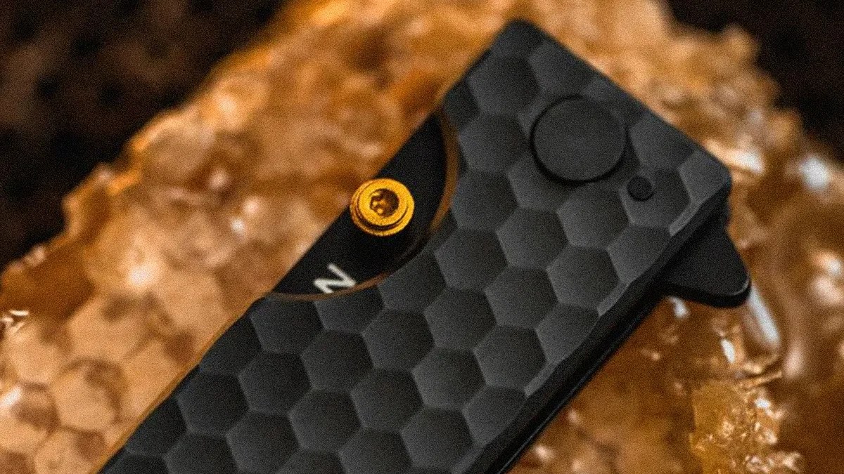 Black textured folding knife with hexagonal pattern and gold screw on a brown crystalline surface.