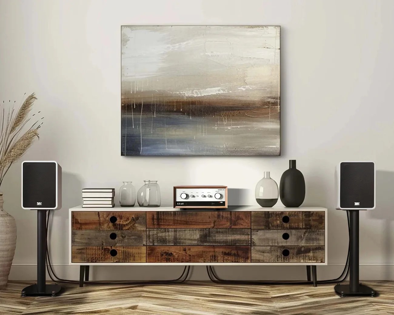 Two black speakers on stands flank a wooden cabinet with a stereo amplifier and decorative vases under an abstract painting.
