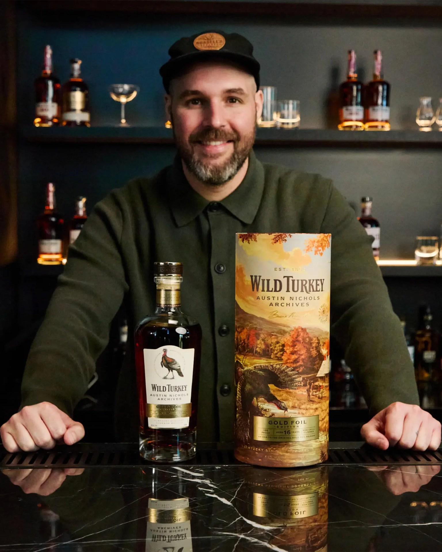 Man in a dark green shirt and black cap standing behind a bottle and box of Wild Turkey Gold Foil bourbon on a black marble counter.