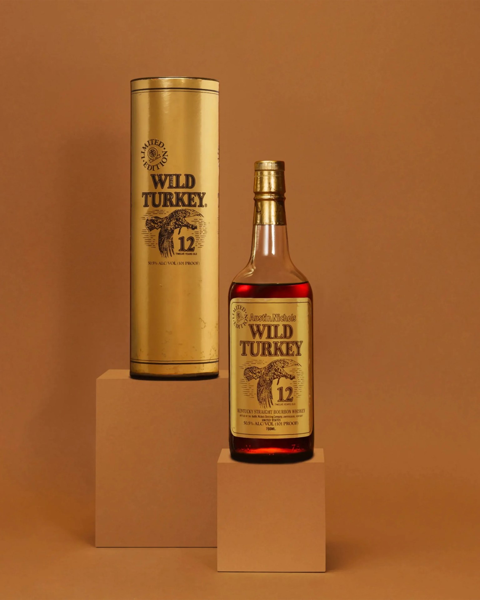 Bottle and gold cylindrical box of Wild Turkey 12-year-old Kentucky Straight Bourbon Whiskey on brown pedestals.