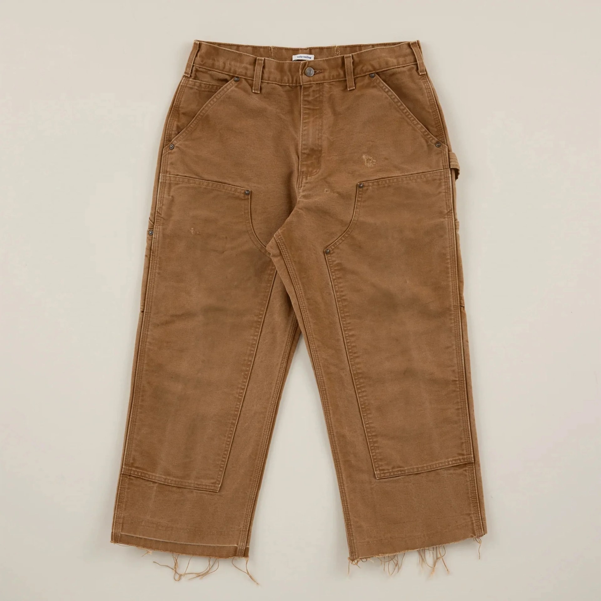 Brown workwear-style pants with large front pockets and frayed hems laid flat on a beige surface.
