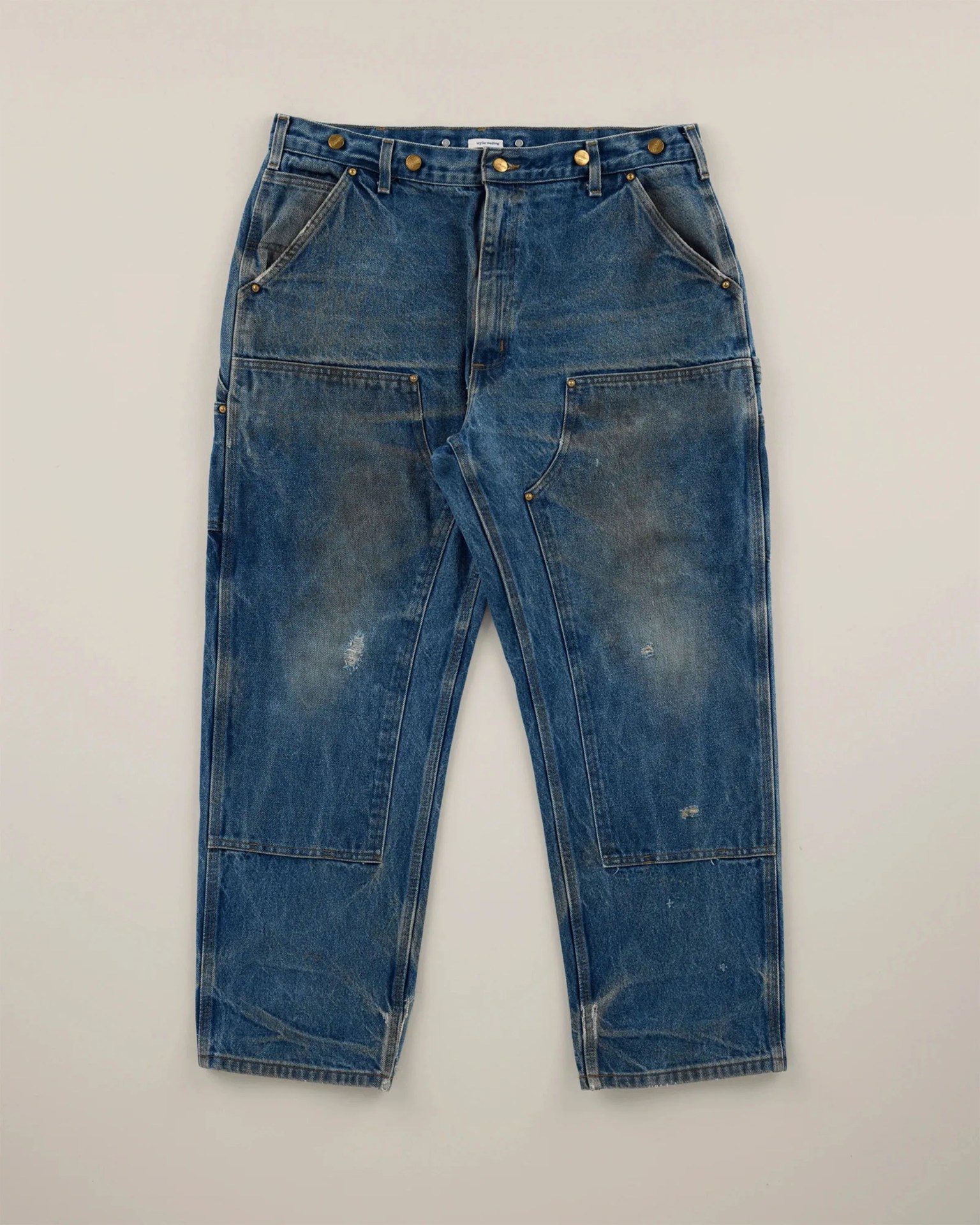 Blue denim carpenter jeans with large front pockets and brass rivets laid flat on a light surface.