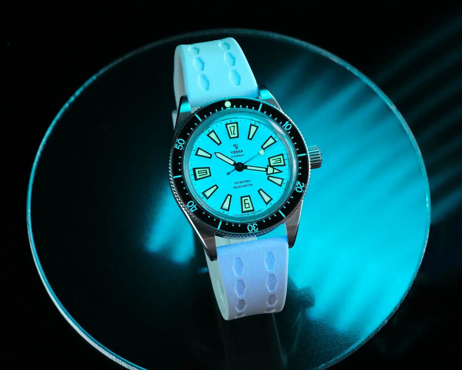 Silver Yema Superman wristwatch with black bezel, luminous blue dial, and white textured strap on a reflective blue surface.