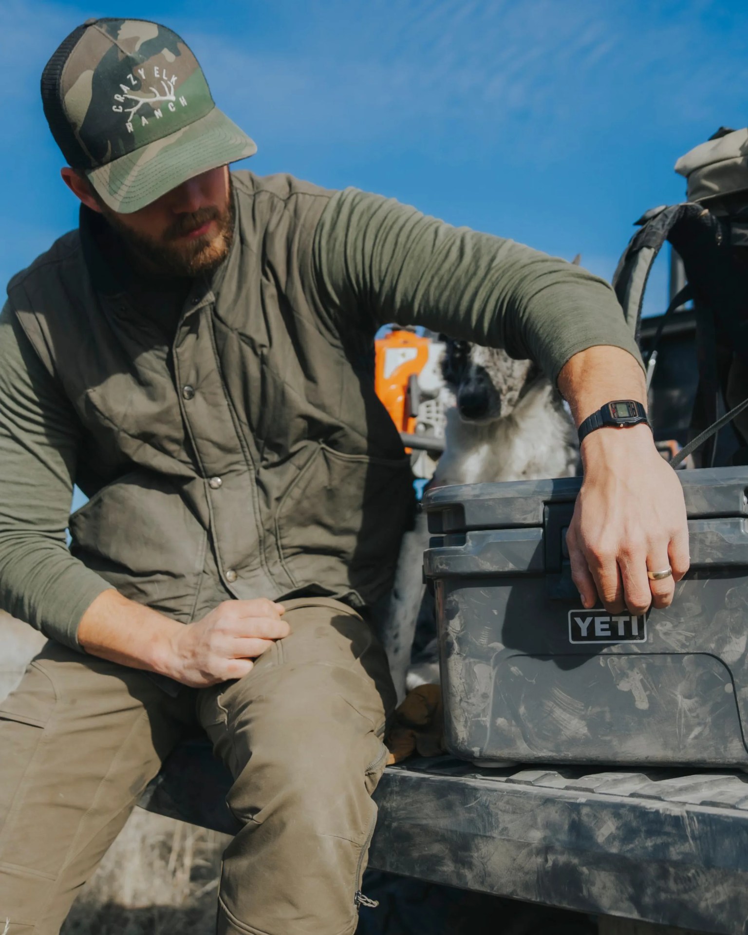Man wearing a camouflage Crazy Elk Ranch cap and green jacket sitting beside a gray YETI cooler with a dog in the background.