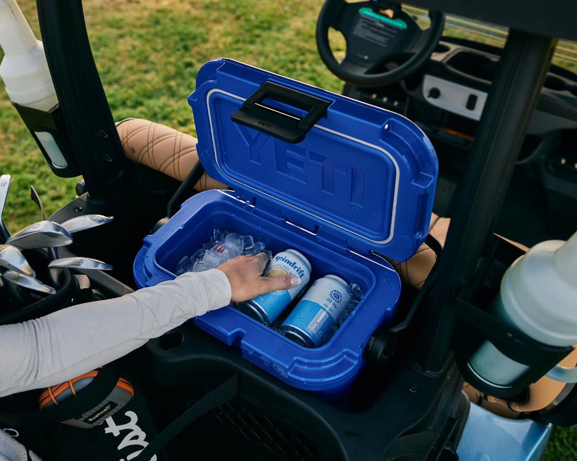 Blue YETI cooler with ice and cans inside, placed on a golf cart seat with a hand reaching for a can.