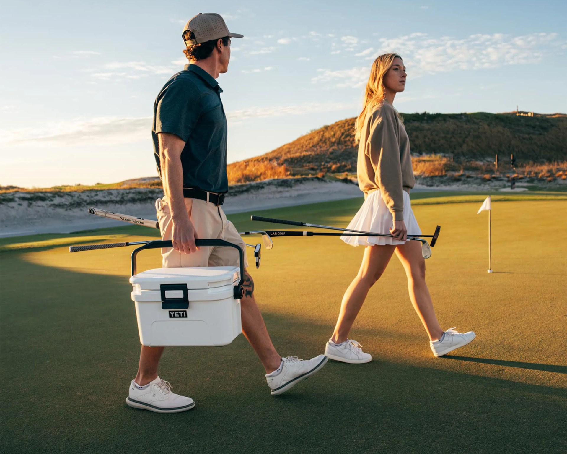 Two people walking on a golf course at sunset, one carrying a white YETI cooler and golf clubs.