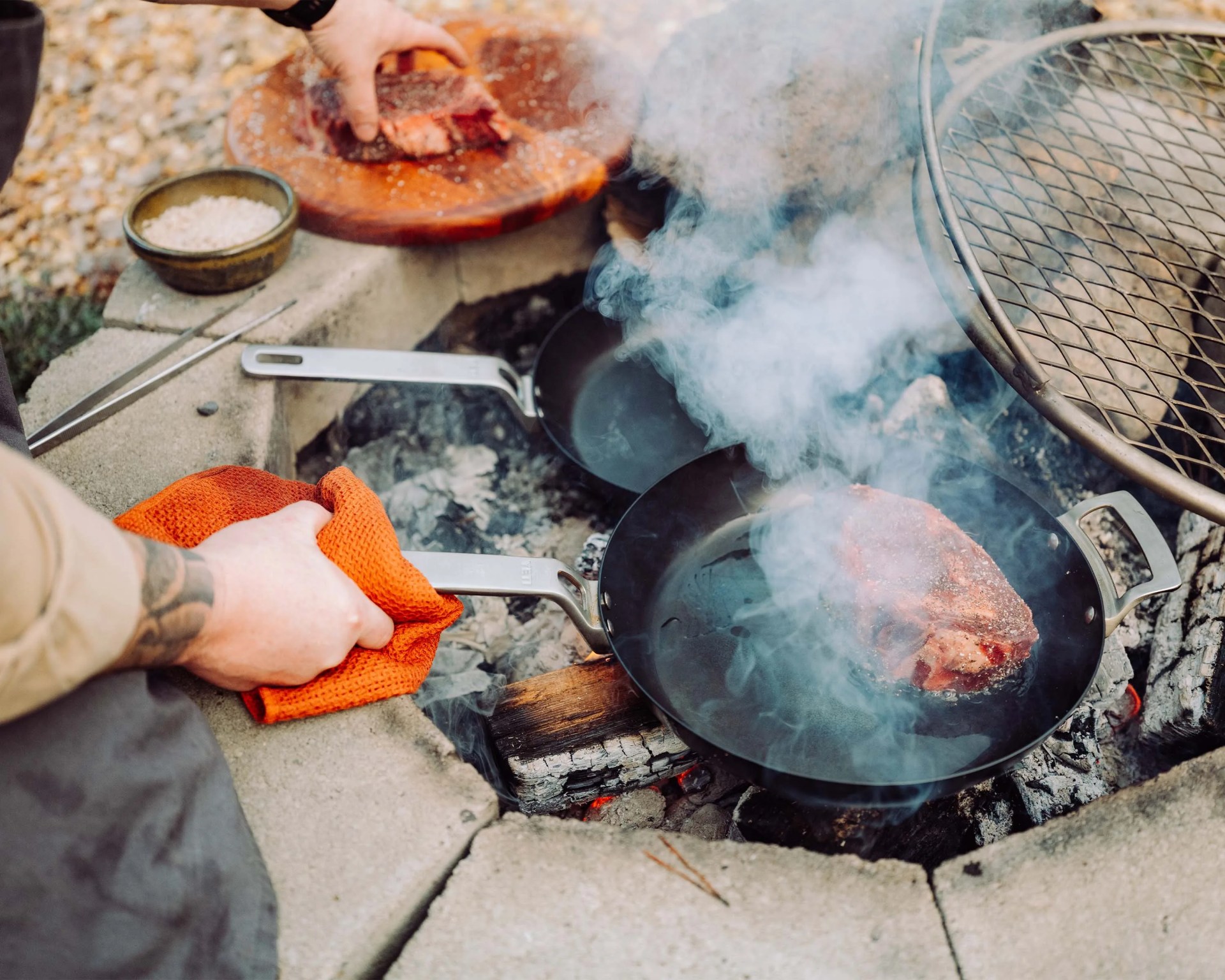 Person holding a black frying pan with steak cooking over an outdoor fire pit with smoke rising.