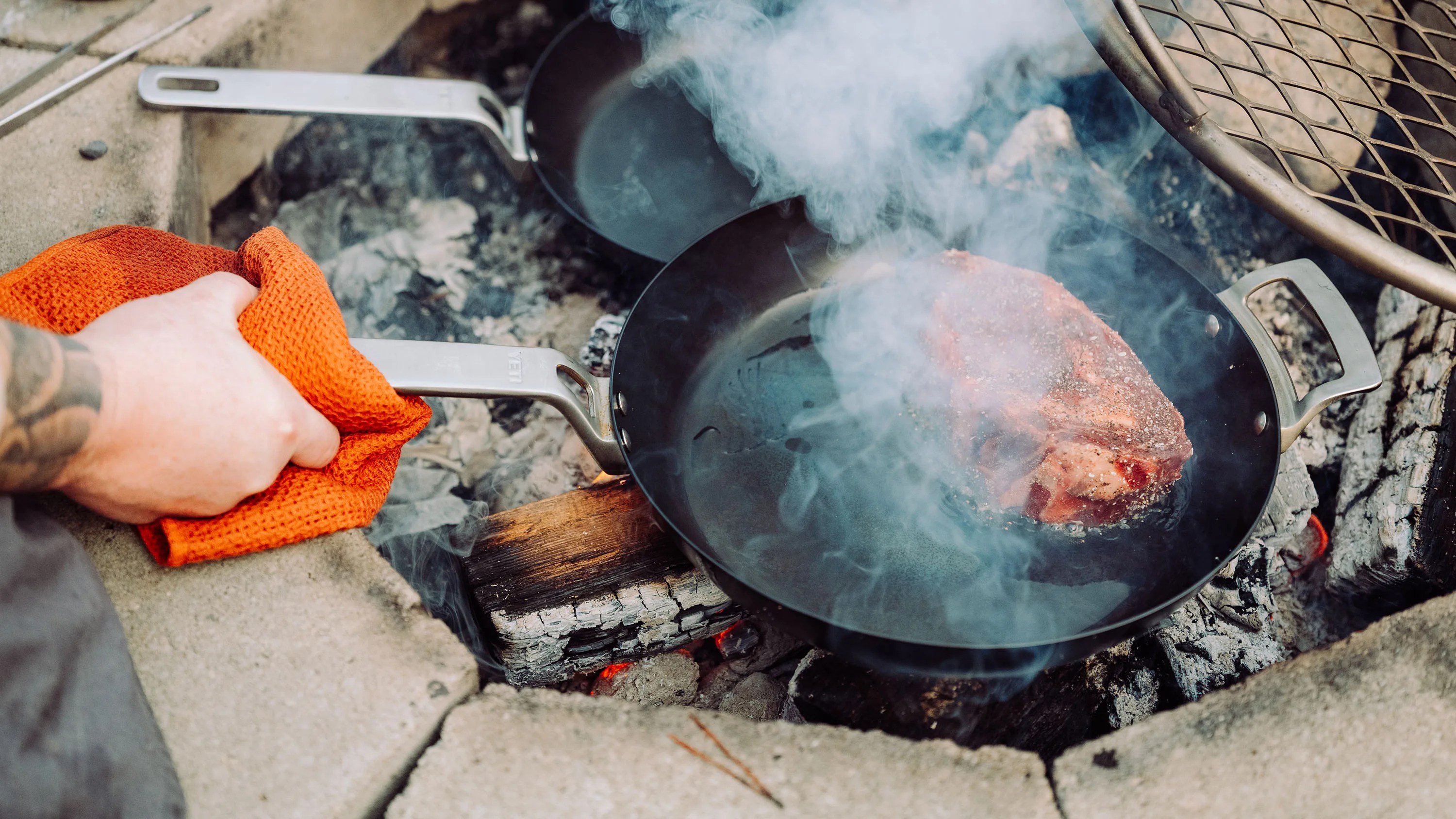 Hand holding an orange cloth gripping the handle of a smoking black frying pan with steak cooking over a wood fire.