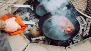 Hand holding an orange cloth gripping the handle of a smoking black frying pan with steak cooking over a wood fire.