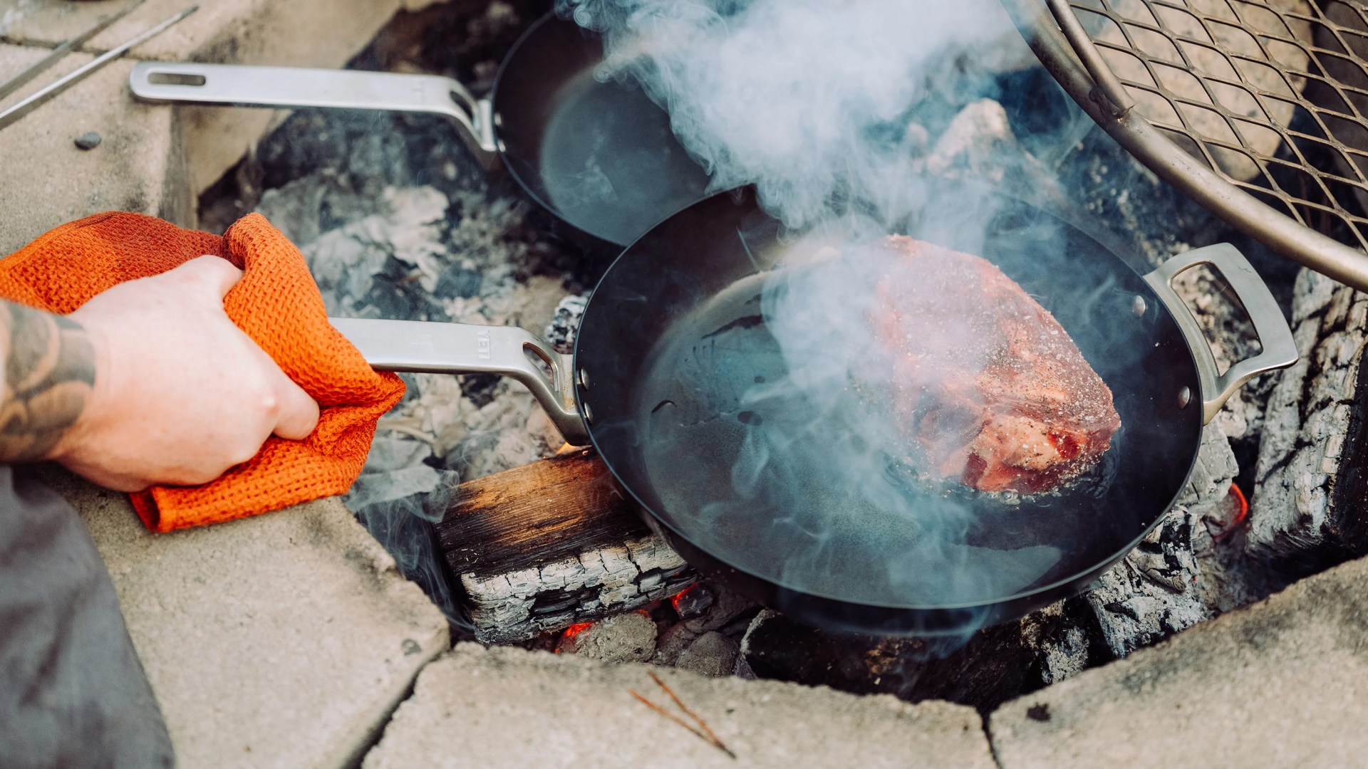 Hand holding an orange cloth gripping the handle of a smoking black frying pan with steak cooking over a wood fire.